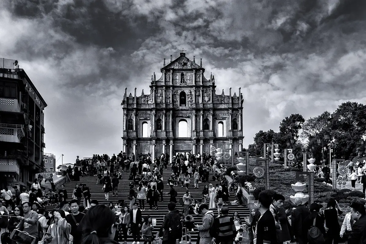 The carved stone facade of the Ruins of St Paul's at the top of a wide staircase crowded with visitors, under a dark cloudy sky, in black and white