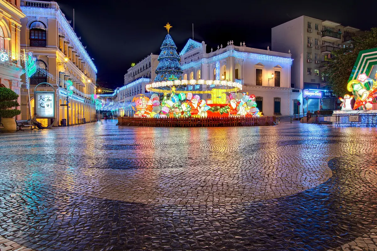 Senado Square at night, with wave-patterned stone paving, a lit Christmas tree and colourful lanterns, and arcaded buildings on both sides